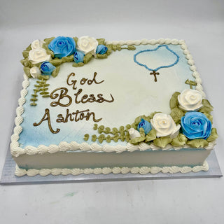 A rectangular cake inspired by the Blessed Necklace, decorated with blue and white roses, green leaves, and a beaded necklace with a gold cross. “God Bless Ashton” is written in brown icing.