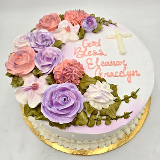 The Flower Goddess is a round white cake adorned with pastel pink, purple, and white buttercream flowers, green leaves, a white cross, and "God Bless Eleanor Gracelyn" written in pink frosting.
