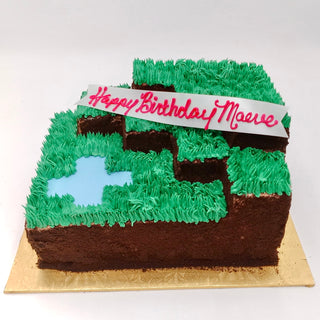 A square cake called "Minecraft Grass," decorated to look like a Minecraft landscape with green icing, blue "water," brown cake “dirt,” and a pink-lettered "Happy Birthday Maeve" banner—perfect for kids and Minecraft fans.