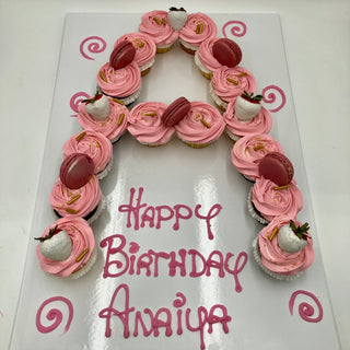 The Letter Cupcakes cake is shaped like an "A" and decorated with pink frosting, macarons, and dipped strawberries. "Happy Birthday Anaiya" is written in pink icing on the white board beneath the cake.
