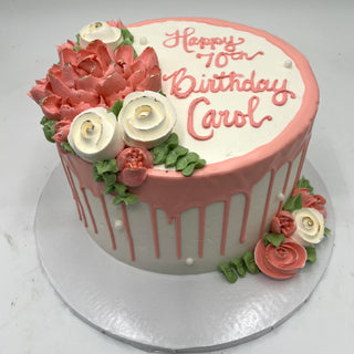 Dripped Beauty: A round white custom cake with pink dripped icing, adorned with pink and white buttercream flowers. Topped with a plaque reading "Happy 70th Birthday Carol" in pink—ideal for specialty birthday celebrations.