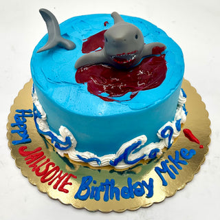 The Shark Attack cake is a round blue birthday cake with a shark figurine emerging from red “blood” icing. “Happy JAWSOME Birthday Mike!” is written in blue and red icing on a gold board.