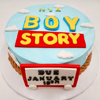 Boy Story round cake styled like the Toy Story logo, featuring "Its a Boy Story" on top and "Due January 19th" on a red and white sign. Decorated with blue sky, white clouds, and brown edging for a playful finish.