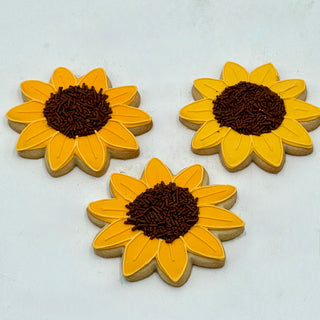 Three Sunflower Cookies with yellow icing petals and chocolate sprinkle centers are arranged on a white surface.