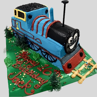 A vibrant Thomas 3D cake, designed as Thomas the Tank Engine on green icing with trees and tracks, features "Happy 2nd Birthday Jack" in red icing on the birthday cake board.
