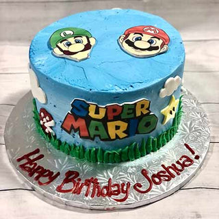 A custom kids cake decorated with blue icing, Luigi and Mario faces, Super Mario text and themed decorations, with "Happy Birthday Joshua!" in red on a silver board. Product name: Mario Logo.