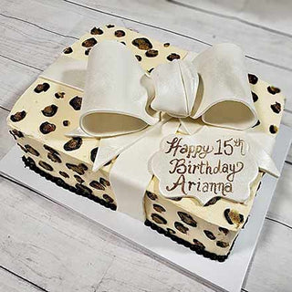 The Cheetah Present is a rectangular custom birthday cake with cheetah-print spots, a big white fondant bow, and a plaque reading "Happy 15th Birthday Arianna" in cursive—one of our Specialty Birthday Designs.