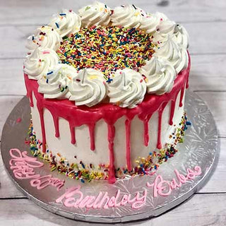 Sprinkle Drip: A round custom birthday cake with white frosting, pink Sprinkle Drip icing, colorful sprinkles, whipped cream swirls, and “Happy Birthday” in pink icing on a silver board—ideal for specialty birthday designs.