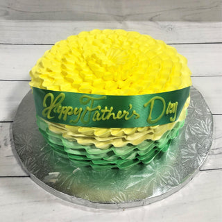 Ruffled Colors is a round cake with yellow and green ombre ruffled frosting, topped with a green ribbon that says "Happy Father's Day" and set on a silver cake board. Contact us for custom pricing on specialty birthday cakes.