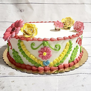 The Fiesta Florals cake features colorful icing flowers and green leaves on white frosting, with pink, yellow, and red designs. This Specialty Birthday Design is beautifully presented on a gold board.