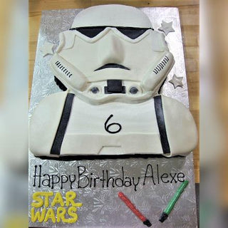 A Star Wars Trooper birthday cake shaped like a stormtrooper helmet with a number 6, "Happy Birthday Alexe" on the board, Star Wars designs, two lightsabers, and fun characters—perfect for kids.