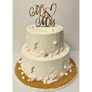 Beach Bride is a two-tier white bridal cake adorned with seashells, pearls, coral designs, and a sandy base. Topped with a gold “Mr & Mrs” heart topper, it's the perfect beach-themed wedding centerpiece.