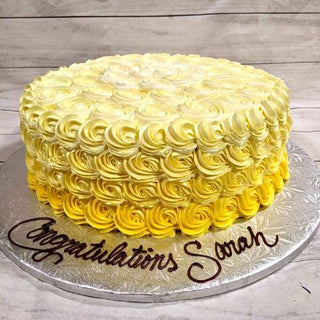 The Rosette Layer specialty cake, topped with yellow and white rosette frosting and “Congratulations Sarah” in brown icing, is beautifully presented on a silver board.