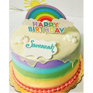 The Rainbow Drip is a round specialty birthday cake featuring white icing drips, “Happy Birthday,” sun decorations, a custom name plaque, and pink piping at the base—ideal as a custom cake for any celebration!.