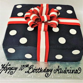 The Gift Box is a square navy blue cake with white polka dots, red and white ribbon, and a bow. Ideal for specialty birthdays, personalized with “Happy 10th Birthday Audrina” in black icing. Custom pricing available.