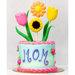 A round pastel blue cake with "MOM" in purple icing, pink borders, and three vibrant Floral Pops—two tulips and a daisy—on top. Perfect for Specialty Birthday Designs; ask about Floral Pops custom cake pricing for your celebration!.