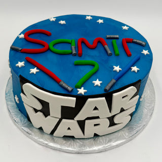 A round blue cake featuring the Star Wars Logo on the front, decorated with white star accents, Samir’s name, a number 7, and colored icing lightsabers on top.