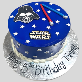 Star Wars Sky is a blue custom kids cake featuring Darth Vader’s mask, two lightsabers, and white stars. “Happy 5th Birthday Tommy” is written on the silver base, perfect for a fun birthday celebration.