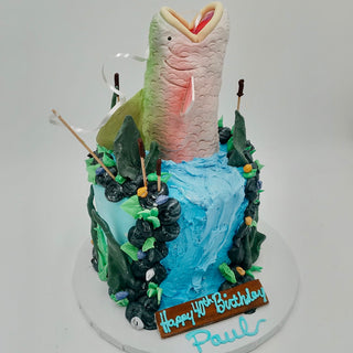 The Fishing Bass birthday cake features a waterfall design with a large fish leaping out, edible rocks, plants, cattails, and a plaque reading "Happy 47th Birthday Paul" in blue icing.