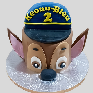 A round cake decorated as a cartoon animal face with big eyes, large ears, and a blue Paw Patrol Hat with "Keonu-Bleu 2" in yellow letters, perfect for kids and presented on a silver patterned cake board.