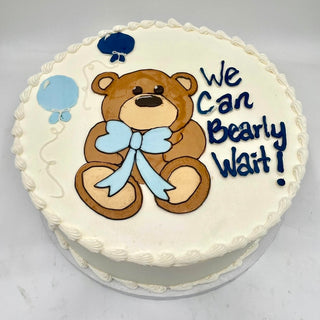 The Bearly Wait cake is round and white, featuring a cute teddy bear with a blue bow, two blue balloons, and “We Can Bearly Wait!” written in blue icing.