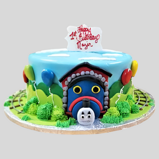 The Thomas Fondant is a round custom kids cake with a Thomas face, colorful balloons, green bushes, train track details, and a white plaque that says "Happy 1st Birthday Najin" in red icing.