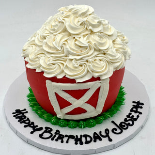 The Barn Smash is a round cake designed as a red barn, featuring white frosting doors and a swirled white icing roof. "Happy Birthday Joseph" is written in black icing on the cake board.