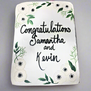 The Flower Vines cake is a rectangular cake decorated with white flowers, green leaves, and engagement flower vines, featuring “Congratulations Samantha and Kevin” written in black icing at the center.