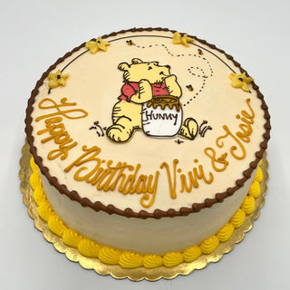 The "Winnie Drawn" birthday cake features Winnie the Pooh eating from a “hunny” pot, yellow flowers, brown piping, and yellow icing borders. Cursive text says “Happy Birthday Vivi & Josie.”.