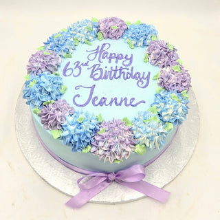 A round blue Hydrangea Edging cake topped with purple, blue, and white flowers, a purple ribbon, and "Happy 63rd Birthday Jeanne" written in purple icing. The cake is displayed on a silver board.