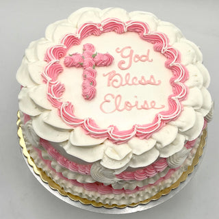 Holy Draping: round white cake with pink and white frosting, featuring a piped cross and "God Bless Eloise" on top, scalloped borders, set on a gold-edged cake board.