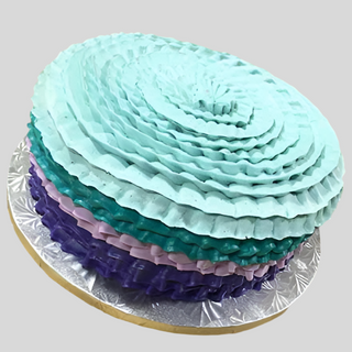 The Ruffled Ombre is a round custom birthday cake with teal, light blue, lavender, and dark purple ruffled ombre frosting on a silver foil board—ideal for specialty birthday designs.