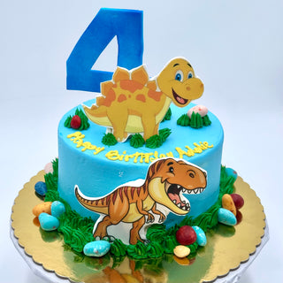 The Dinosaur Cartoon cake features blue icing, cartoon dinosaurs, a large blue number 4 topper, colorful candy eggs, and "Happy Birthday Addie" in yellow icing on a gold board.
