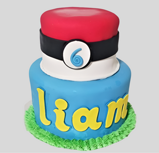 A two-tiered custom Pokemon Name cake features a Poké Ball top, blue lower tier with Liam’s name in yellow, green icing grass at the base, and a “6” centerpiece—ideal for a kids’ birthday celebration.