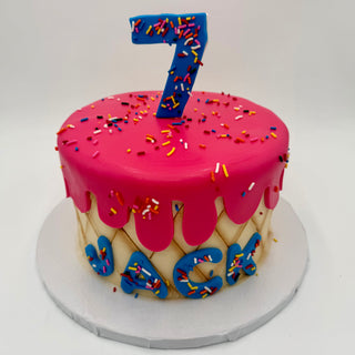 The Ice Cream Drip cake features pink icing, colorful sprinkles, a large blue number 7 topper, diamond-patterned sides, and "Jack" in blue letters. It sits on a white board.