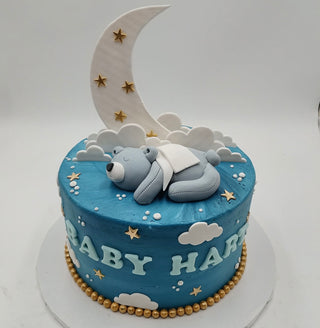 Bear Baby features a blue cake decorated with “BABY HART,” gold stars, clouds, a crescent moon, and a fondant bear sleeping under a white blanket on a white board with gold bead trim at the base.