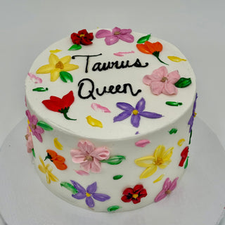 A round white cake called Flower Queen, decorated with colorful buttercream flowers and "Flower Queen" written on top in black icing.