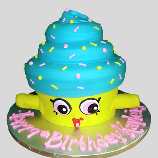 The Shopkins Cupcake is a custom cartoon-style cake with blue frosting, colorful sprinkles, big eyes, and a smile. “Happy Birthday Katelyn” is piped in pink icing on the silver board. A fun kids’ birthday treat!.