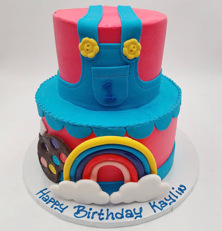 Ms. Rachel Rainbow is a two-tiered birthday cake with colorful fondant featuring blue overalls, a rainbow, clouds, a paint palette, and the number 1. “Happy Birthday Kaylin” is written on the cake board.