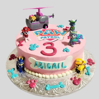 The Paw Patrol Party cake features figurines, a helicopter, paw prints, and bones with blue and pink accents. It displays the number 3 and Abigail's name—perfect for a themed birthday celebration!.