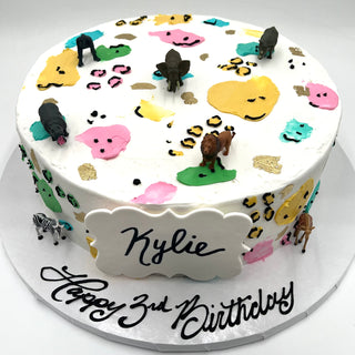 Round white birthday cake decorated with colorful animal print and Safari Figures. A plaque reads Kylie, and the cake board says "Happy 3rd Birthday" in black script.