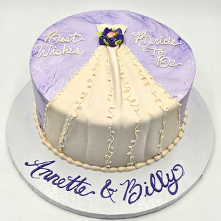 The Wedding Dress cake features purple and white icing, says "Best Wishes Bride To Be" on top, and has "Annette & Billy" in purple script at the base—a perfect choice for engagement celebrations.