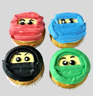 Ninjago Cupcakes: Four cupcakes topped with colorful ninja faces—each with a red, blue, black, or green mask and expressive frosted eyes. These custom treats are perfect for kids' parties.
