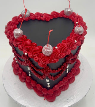 Black Heart: A heart-shaped cake with smooth black icing, topped with red piped frosting, silver balls, and glittery faux cherries, served on a plain white round base.