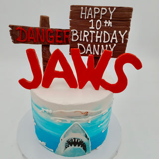 The Jaws Bite cake features a shark from Jaws, a swimmer, water, red "JAWS" letters on top, plus DANGER and HAPPY 10th BIRTHDAY DANNY signs.
