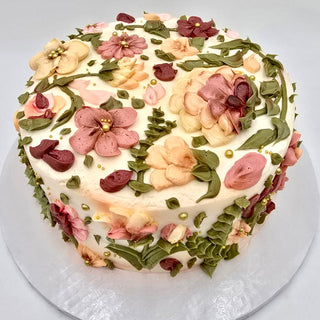 Elaborate Florals: A round cake adorned with pink, peach, and cream buttercream flowers, green leaves, and gold pearls on a white background.