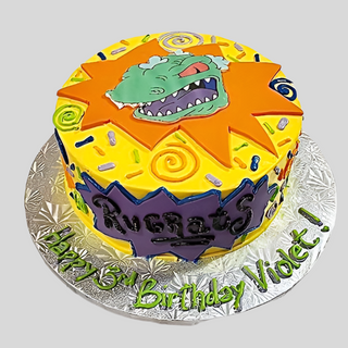 A Rugrats Retro kids cake with the Rugrats logo and cartoon dinosaur face, designed in yellow, orange, and purple. The silver cake board says “Happy 3rd Birthday Violet!” in green icing.