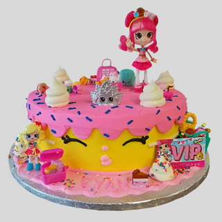 The Shimmer Face cake is round with pink icing, sprinkles, toy figurines, whipped cream shapes, and a cheerful Shimmer Face design. On top are a doll figure and Kids Characters, plus VIP and custom pricing decorations around the base.