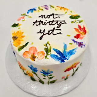 The Artistic Garden is a round frosted cake adorned with colorful painted flowers and leaves, topped with "non-thirty yet" handwritten in chocolate icing.