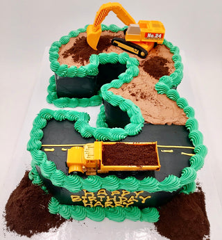Number Trucks is a birthday cake shaped like the number 3, decorated with green icing, toy construction vehicles, chocolate "dirt" crumbs, a road design, and "Happy Birthday" for a fun construction theme.